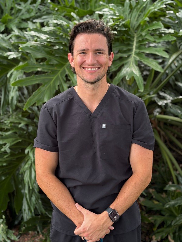 Portrait of Dr. Samuel Pulsipher smiling in charcoal gray medical scrubs against a leafy green plant background