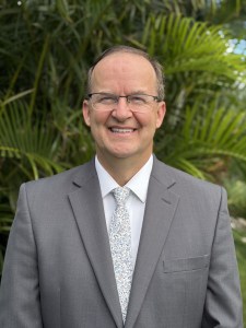 Professional headshot of Dr. Michael Fowler wearing eyeglasses and a gray suit with a patterned tie against green foliage