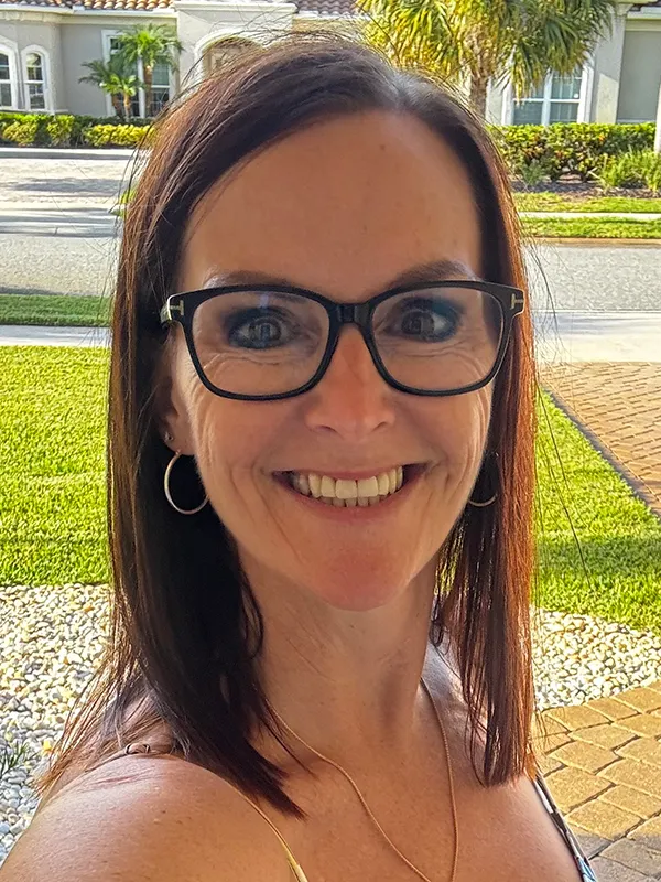 Headshot of a smiling woman with dark straight hair and black eyeglasses, outdoors on a paved walkway