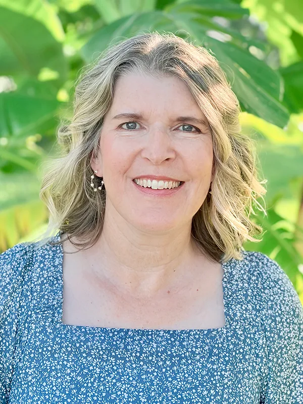 Headshot of a smiling woman with wavy blonde hair wearing a blue patterned top against a lush green leafy background