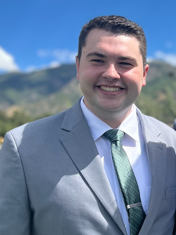 Portrait of a smiling man in a light gray suit, white shirt, and green patterned tie with a scenic mountain backdrop